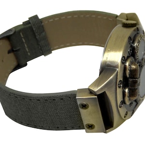 K&ouml;nnte beinhalten: Eine bronzefarbene Uhr mit einem einzigartigen, mehrschichtigen Design. Die Uhr hat ein graues Canvas-Armband mit N&auml;hten.