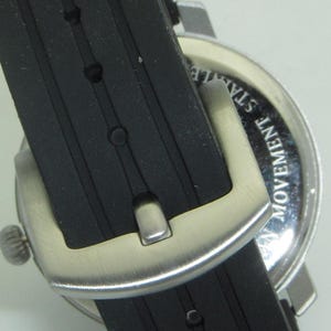 May include: Close-up of a black rubber watch band with a silver buckle. The watch face is visible in the background and says "Movement Stainless".