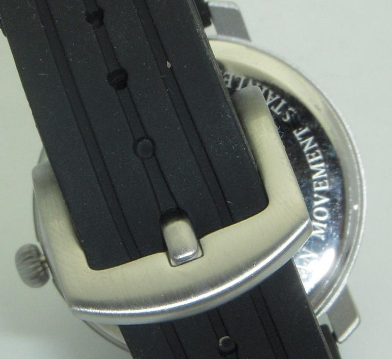 May include: Close-up of a black rubber watch band with a silver buckle. The watch face is visible in the background and says "Movement Stainless".