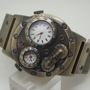 K&ouml;nnte beinhalten: Eine Armbanduhr im Steampunk-Stil mit einem messingfarbenen Geh&auml;use und einem gr&uuml;nen Canvas-Armband. Das Zifferblatt der Uhr hat drei Zifferbl&auml;tter, eines mit r&ouml;mischen Ziffern und zwei mit arabischen Ziffern.