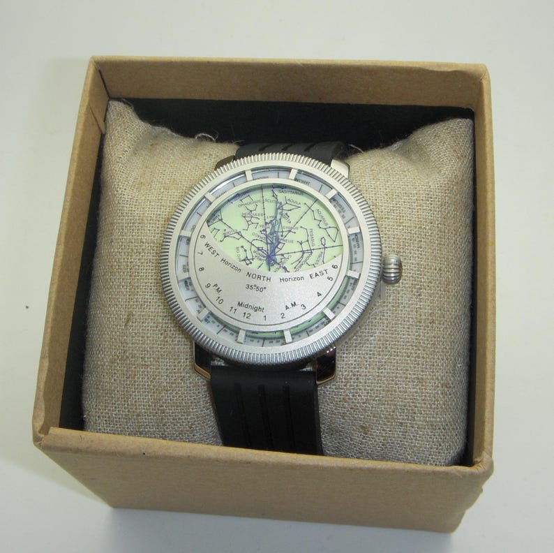 May include: A silver watch with a black band. The watch face features a map of the world with the text "NORTH 35&deg;50'" and "WEST Horizon" and "EAST Horizon".