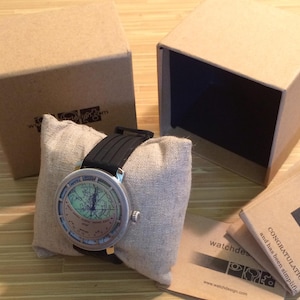 May include: A silver watch with a black band and a world map face. The watch is in a brown cardboard box with a white pillow inside. The box has the text "watchdesign.com" printed on it.