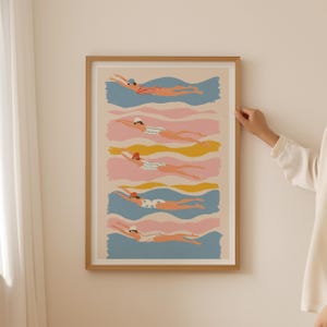 May include: A framed art print featuring six swimmers in various swimsuits and swim caps, each in a different color scheme. The swimmers are depicted in a streamlined position, swimming through wavy bands of blue, pink, and yellow.