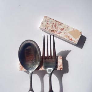 May include: A stainless steel spoon and fork rest on a rectangular terrazzo tile. A second terrazzo tile is positioned above. The spoon and fork are placed on a white surface. The terrazzo tiles have a mix of colors including pink, beige, and white.
