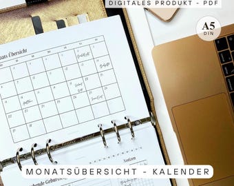 May include: A gold Filofax planner with a calendar page open, showing a monthly overview. The text "DIGITALES PRODUKT - PDF" and "A5 DIN" are visible. A laptop and smartphone are in the background. The calendar is labeled "MONATSÜBERSICHT - KALENDER".