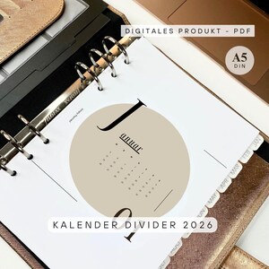 Könnte beinhalten: A5-Planereinlage mit einem Kalender für Januar 2026. Die Einlage zeigt einen beigen Kreis mit dem Monatsnamen und Daten in minimalistischer Schrift. Der offene Planer zeigt Metallringe und beschriftete Register.