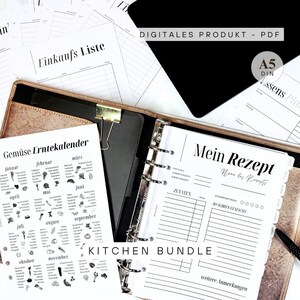 Könnte beinhalten: Eine Sammlung druckbarer Küchenblätter, darunter eine Einkaufsliste, Rezeptseiten und ein Gemüseerntekalender. Die Seiten sind in Schwarzweiß gehalten und werden in einem gold-schwarzen Ordner aufbewahrt. Der Text "KITCHEN BUNDLE" ist sichtbar.