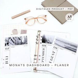 Könnte beinhalten: Ein Planer mit einem transparenten Ordner, der geöffnet ist, um monatliche Dashboard-Seiten anzuzeigen. Die Seiten zeigen Schwarz-Weiß-Fotografien und Kalenderdaten. Der Text "DIGITALES PRODUKT - PDF" und "MONATS DASHBOARD - PLANER" sind sichtbar. Ebenfalls enthalten sind eine Brille, ein Stift und ein Notizbuch.