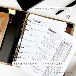 Peut inclure: Un planificateur doré et noir avec des inserts en papier blanc. Les inserts comportent les mots "Essens Plan" et une mise en page de plan de repas hebdomadaire. Le planificateur est ouvert, révélant les pages et une pince dorée.