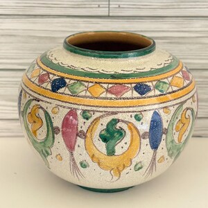 May include: A round ceramic vase with a speckled cream base, featuring a hand-painted decorative band of yellow, green, blue, and pink geometric shapes and floral-like motifs. The vase has a wide opening and a smooth, glazed finish.