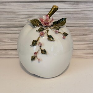 May include: A white ceramic decorative apple with a gold stem and pink floral accents. The apple-shaped container features detailed green leaves and small pink flowers, creating an elegant, nature-inspired design.