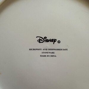 May include: Off-white ceramic plate with the Disney logo in black. The text "MICROWAVE AND DISHWASHER SAFE", "STONEWARE", and "MADE IN CHINA" are printed below the logo. The plate is round.