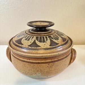 Puede incluir: Una fuente redonda de cerámica con tapa, en tonos beige y marrón. La tapa presenta un diseño decorativo marrón oscuro y un pequeño asa redonda. La fuente tiene dos asas laterales y un acabado moteado.