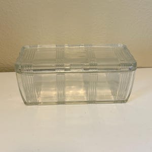 May include: A clear, rectangular glass box with a lid. The box features a grid-like pattern on the lid and vertical lines on the sides. The box is empty and sits on a white surface.