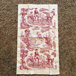 May include: Cream-colored kitchen towel with a red toile de Jouy design featuring historical figures and scenes. The towel has a rectangular shape and includes text at the bottom. The design depicts people in carriages and other historical scenes.
