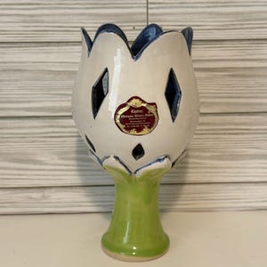May include: A ceramic vase shaped like a tulip with a white upper section featuring diamond-shaped cutouts and a blue rim. The base is a solid light green stem. A red and gold sticker with text is attached to the front of the white section.