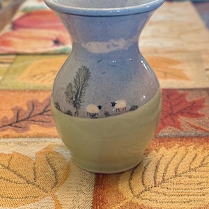 May include: A ceramic vase with a flared rim, featuring a two-tone design. The upper portion is a speckled light blue, while the lower section is a solid pale green. A painted scene depicts two white sheep and a tree in a field.