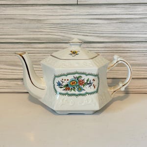 May include: White ceramic teapot with a floral design. The teapot has a gold-trimmed spout, handle, and lid. A rectangular floral pattern with blue, red, and yellow flowers is centered on the teapot's body. The teapot has an octagonal shape.