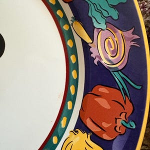 May include: A white ceramic plate with a colorful border featuring illustrations of vegetables. The border is dark blue with red, green, and yellow accents. The vegetables include a red bell pepper, onion, and carrot.