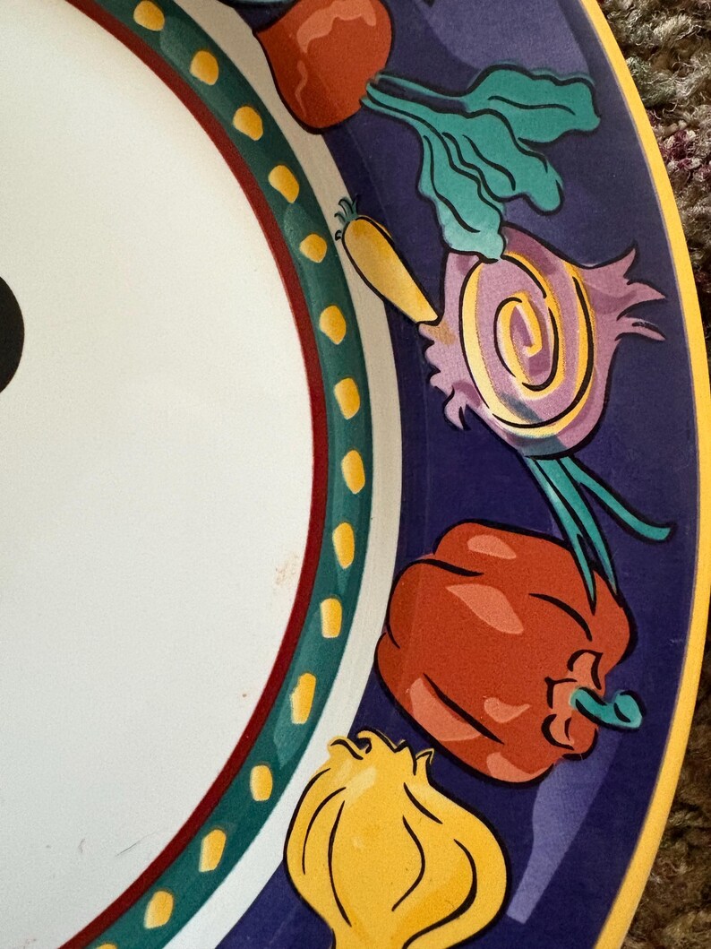 May include: A white ceramic plate with a colorful border featuring illustrations of vegetables. The border is dark blue with red, green, and yellow accents. The vegetables include a red bell pepper, onion, and carrot.