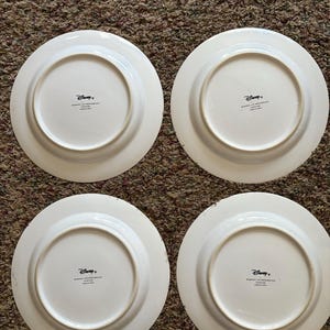 May include: Four white ceramic plates with a circular design. Each plate has a wide rim and a central recessed area. The underside of each plate features the Disney logo and text. The plates are arranged on a brown carpet.