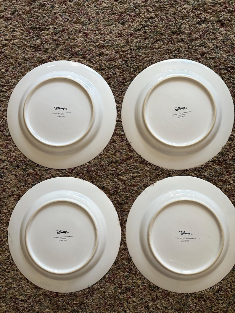 May include: Four white ceramic plates with a circular design. Each plate has a wide rim and a central recessed area. The underside of each plate features the Disney logo and text. The plates are arranged on a brown carpet.