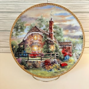 May include: A decorative plate featuring a detailed painting of a cottage scene. The artwork depicts a brick cottage with a smoking chimney, surrounded by lush greenery, colorful flowers, and a wooden fence. The plate has a gold rim.