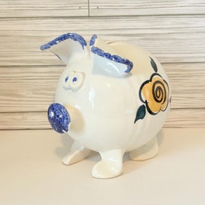 May include: A white ceramic piggy bank with blue accents on the ears, snout, and eyes. The piggy bank features a yellow and black floral design on its side. The bank has four small feet and a slot on top for inserting coins.