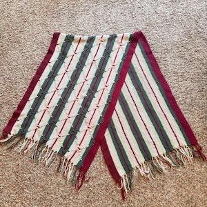 May include: A handmade scarf with a geometric pattern. The scarf features alternating sections of diagonal diamond and vertical stripe designs. Colors include cream, burgundy, and green, with fringe at the ends.
