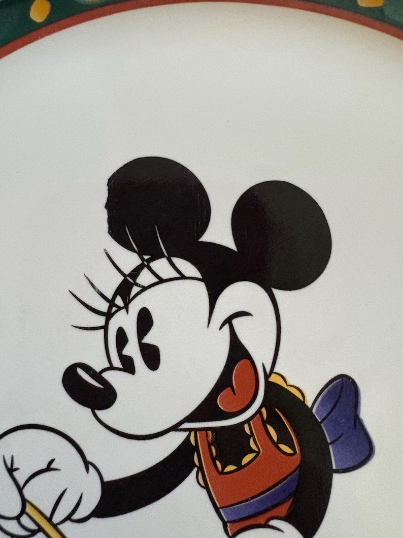 May include: A white plate featuring a smiling Minnie Mouse character. Minnie Mouse is depicted in black, white, red, and orange colors, with a blue bow. The plate has a green and yellow border.