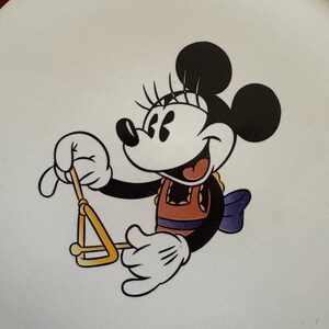 May include: A white plate featuring a cartoon image of Minnie Mouse playing a triangle. Minnie Mouse is wearing an orange top, a purple bow, and white gloves. The plate has a green and yellow border.