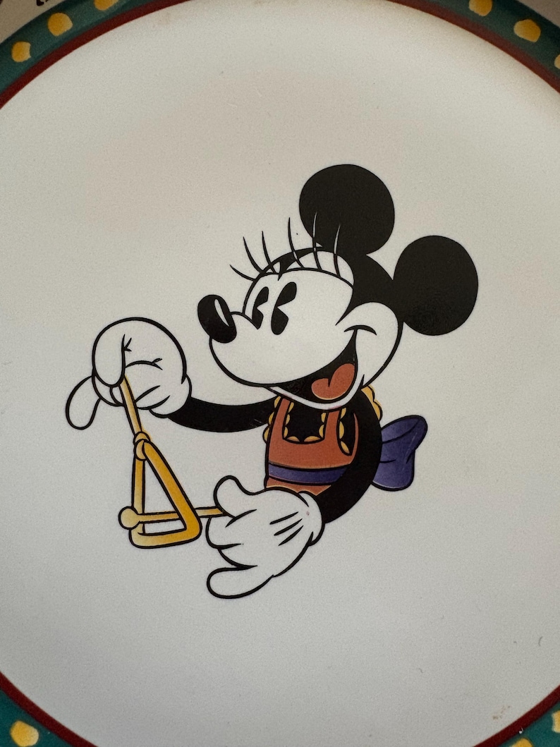 May include: A white plate featuring a cartoon image of Minnie Mouse playing a triangle. Minnie Mouse is wearing an orange top, a purple bow, and white gloves. The plate has a green and yellow border.