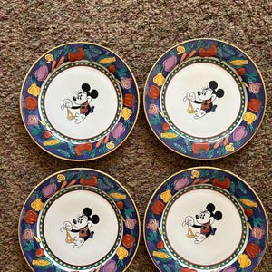 May include: Set of four decorative plates featuring a central image of Mickey Mouse holding scales. The plates have a white center, a blue border with colorful vegetables, and a gold rim. A charming addition to any kitchen.