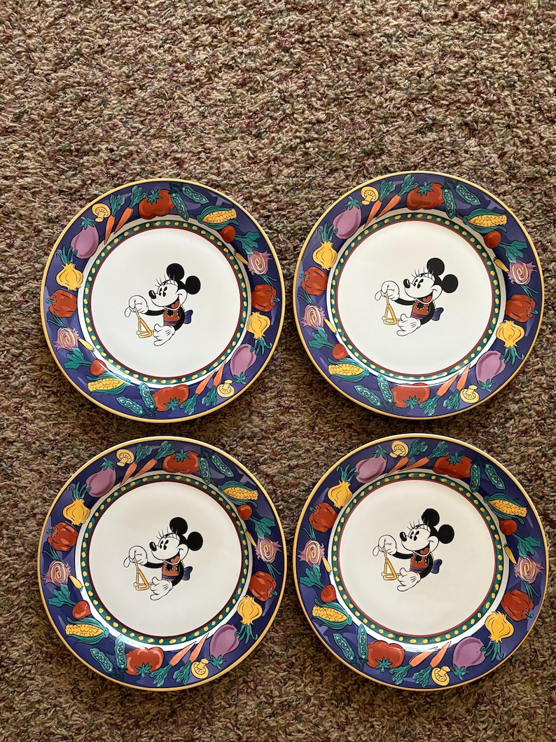 May include: Set of four decorative plates featuring a central image of Mickey Mouse holding scales. The plates have a white center, a blue border with colorful vegetables, and a gold rim. A charming addition to any kitchen.