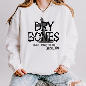 Pu&ograve; includere: Felpa bianca con una grafica nera di una croce con le parole "Dry Bones" e il testo "Hear the Word of the Lord Ezekiel 37:4"