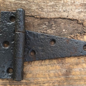 May include: A black, T-shaped hinge with a textured surface and three holes on the horizontal section. The hinge is made of metal and appears aged or rustic. The background is a wooden surface.