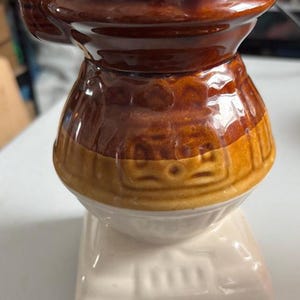 May include: A brown and white ceramic mug shaped like a wood-burning stove. The mug has a brown top and a white base with a brown band around the middle. The mug is decorated with a brown and white design.