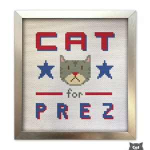 May include: A framed cross-stitch design featuring a gray cat with red nose and blue eyes. The cat is surrounded by two blue stars and the text "CAT for PREZ" in red and blue.