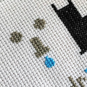 May include: Close-up of a cross-stitch pattern with a black silhouette of a cat, brown and blue accents, and a white background.