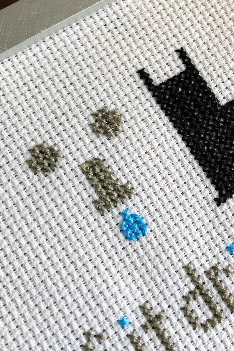 May include: Close-up of a cross-stitch pattern with a black silhouette of a cat, brown and blue accents, and a white background.