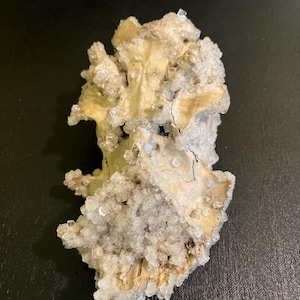 May include: A natural, beige-coloured mineral specimen with clusters of small, clear crystals. The mineral has a rough, textured surface and is set against a dark background. The crystals sparkle, catching the light.