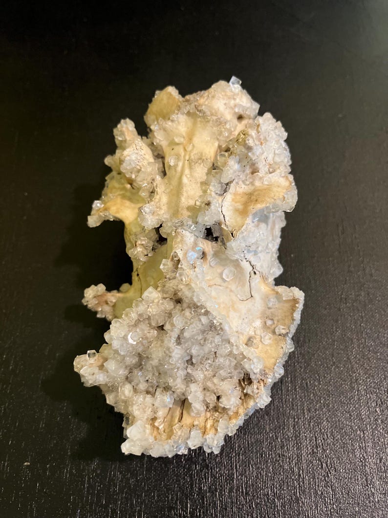 May include: A natural, beige-coloured mineral specimen with clusters of small, clear crystals. The mineral has a rough, textured surface and is set against a dark background. The crystals sparkle, catching the light.