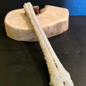 May include: An elongated mineral specimen with a crystalline structure, covered in small, sparkling crystals. It rests on a wooden base. The background is a dark surface with a blue backdrop. The mineral is light-coloured.