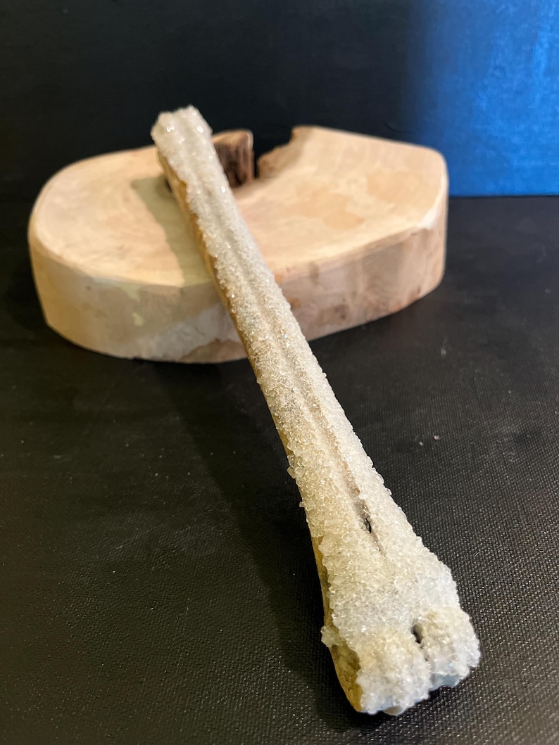 May include: An elongated mineral specimen with a crystalline structure, covered in small, sparkling crystals. It rests on a wooden base. The background is a dark surface with a blue backdrop. The mineral is light-coloured.