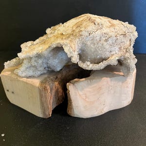 May include: A natural geode crystal formation with a rough, textured surface, resting on a light-coloured, carved wooden base. The crystal features a mix of beige and clear crystalline structures. The wooden base has a natural, unfinished appearance.