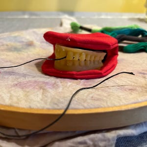 Puede incluir: Una boca de tela roja con un juego de dientes de plástico amarillo en su interior. La boca está en un aro de bordado de madera con hilo negro.