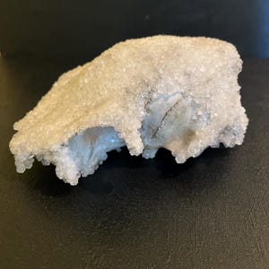 May include: A natural crystal formation with a rough, textured surface. The crystal has a light, frosted appearance with a granular texture. The base of the crystal has a light blue hue. The crystal is set against a dark background.