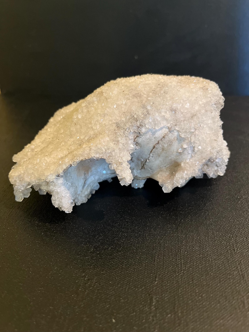 May include: A natural crystal formation with a rough, textured surface. The crystal has a light, frosted appearance with a granular texture. The base of the crystal has a light blue hue. The crystal is set against a dark background.