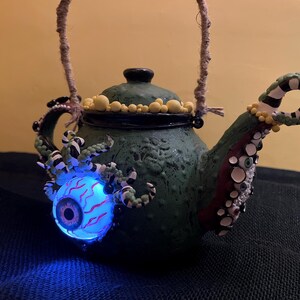 May include: A green ceramic teapot with a rope handle, decorated with tentacles, an illuminated eyeball, and yellow accents. The teapot has a dark green lid and a spout adorned with white and black stripes.