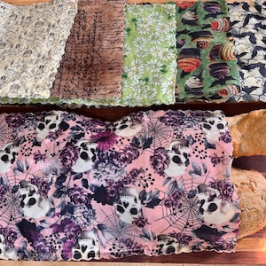 May include: Assortment of reusable food wraps with various patterns, including dogs, script, daisies, cupcakes, bats, and skulls with spiderwebs and flowers. Two loaves of bread are wrapped in a pink skull-themed wrap, showcasing the product's use.